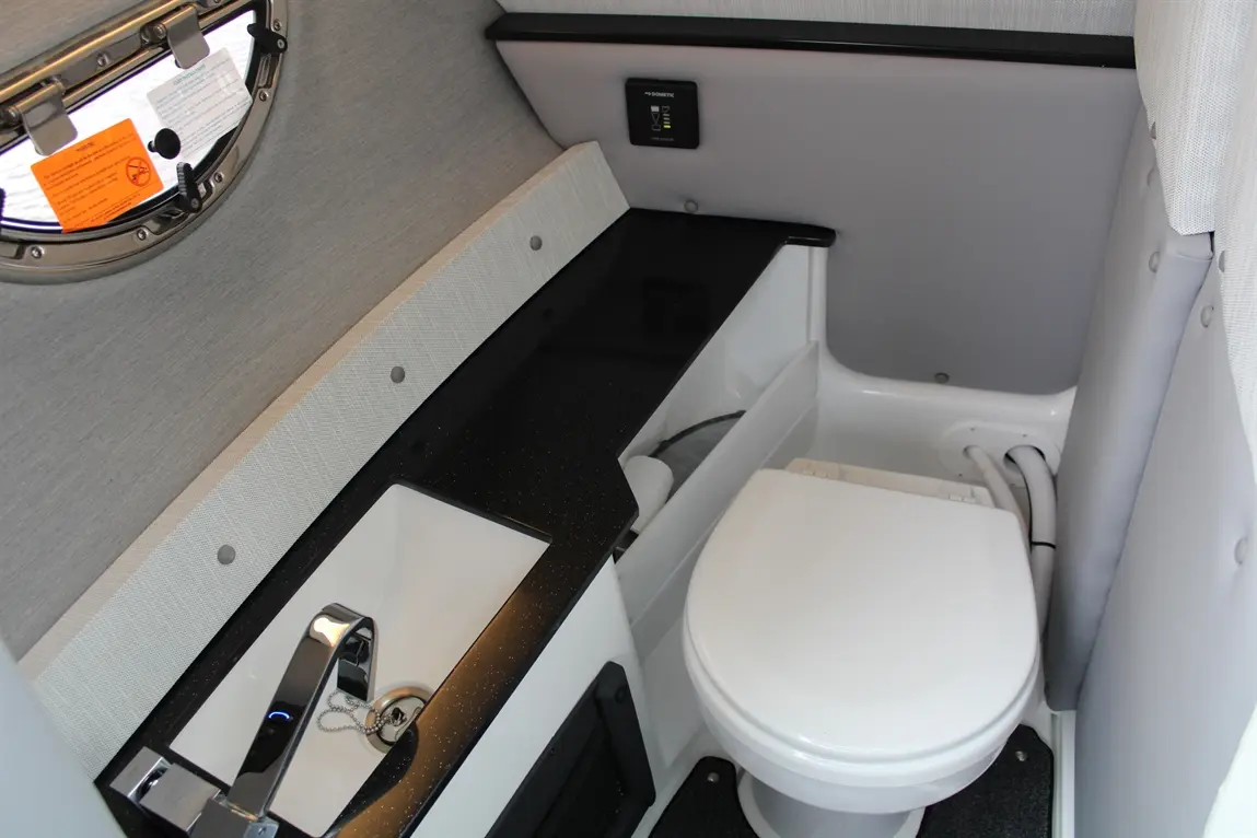 Compact boat bathroom featuring a toilet, sink with faucet, and window with safety notices. Modern design and functionality.